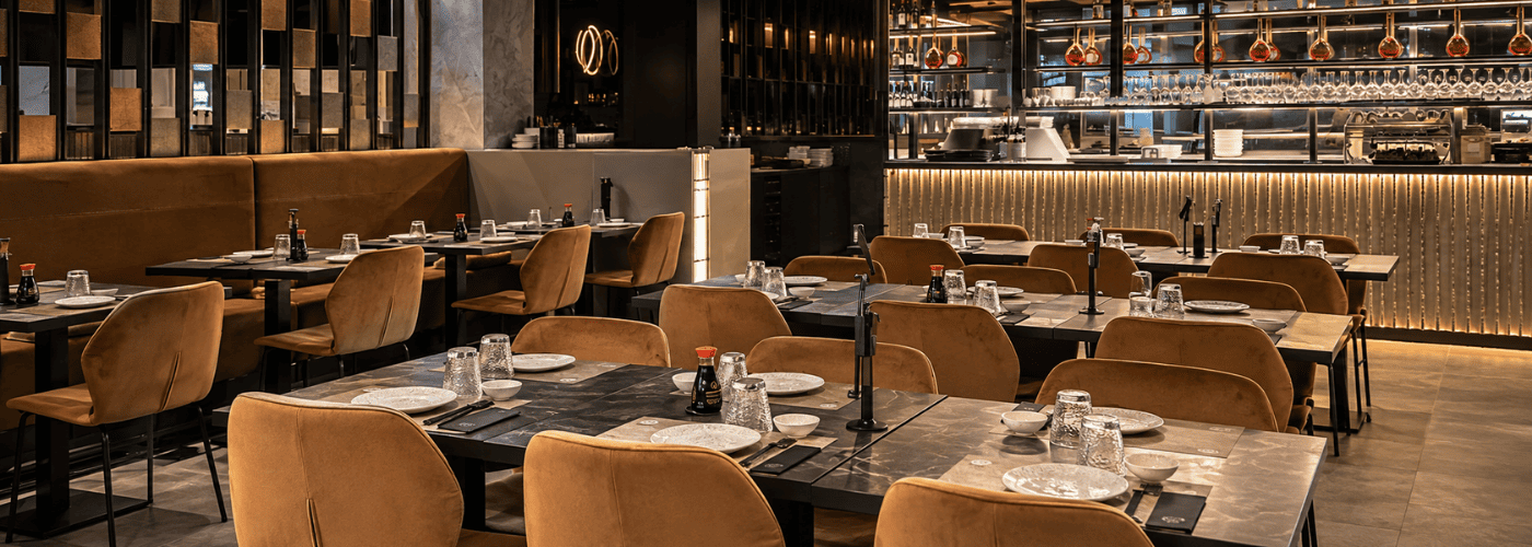 Imagen interior de un restaurante Sushisom con mesas preparadas, decoración contemporánea y zona de servicio al fondo. Refuerza la experiencia de restaurante japonés y el ambiente donde se disfruta el Menú Infinity.
